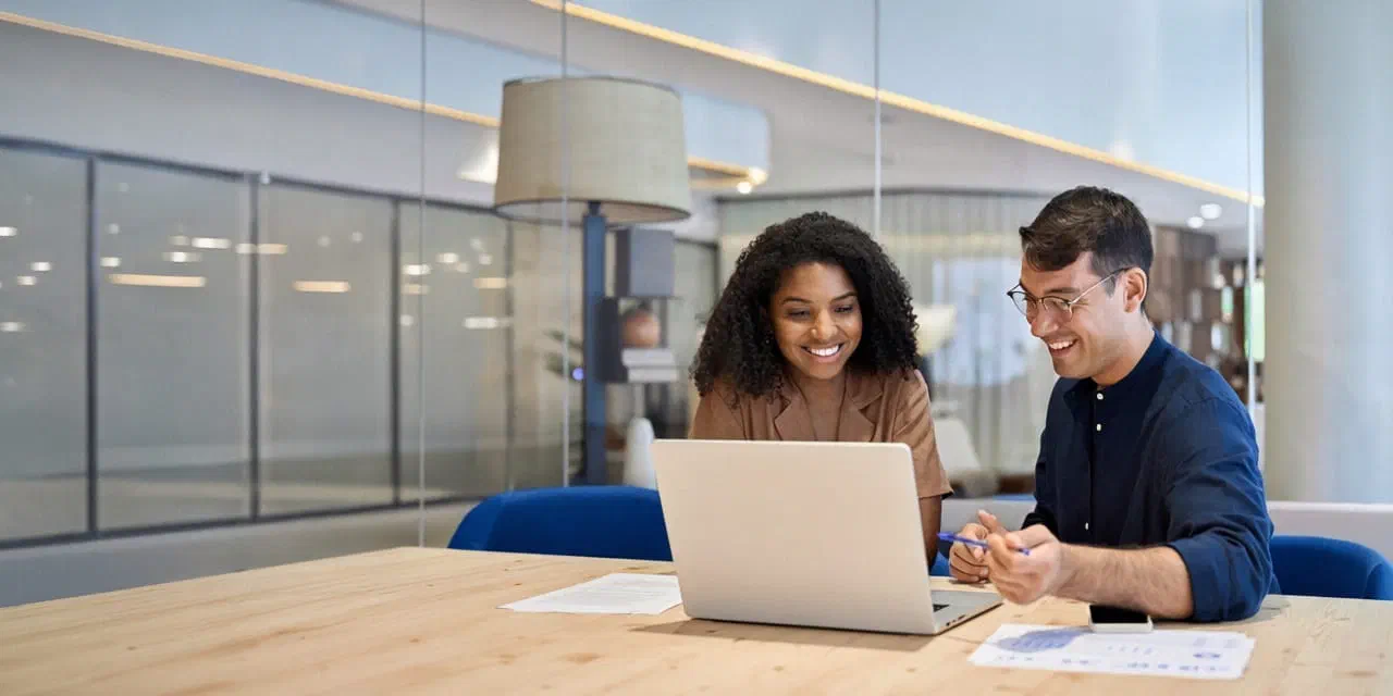 An editorial team of a man and a woman sitting at an office desk, smiling and talking.